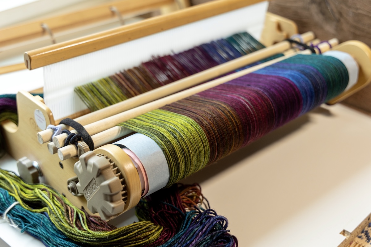 A warp is half-wound onto the loom using a DIY tensioning device consisting of dowels and elastic bands.